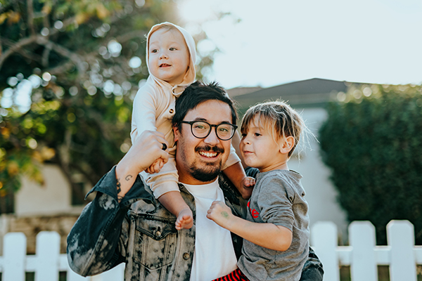 Asian and mixed family outside. Father with two children in front of white picket fence and home smiling.